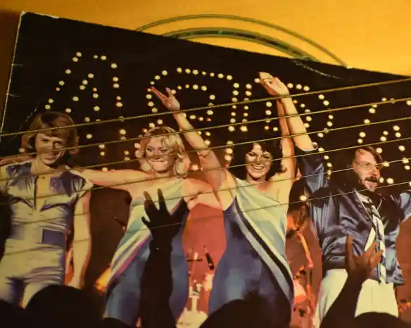 Four performers dressed in vibrant, sequined outfits pose with raised arms on stage, illuminated by bright lights. The crowd's hands can be seen reaching towards them in the foreground.