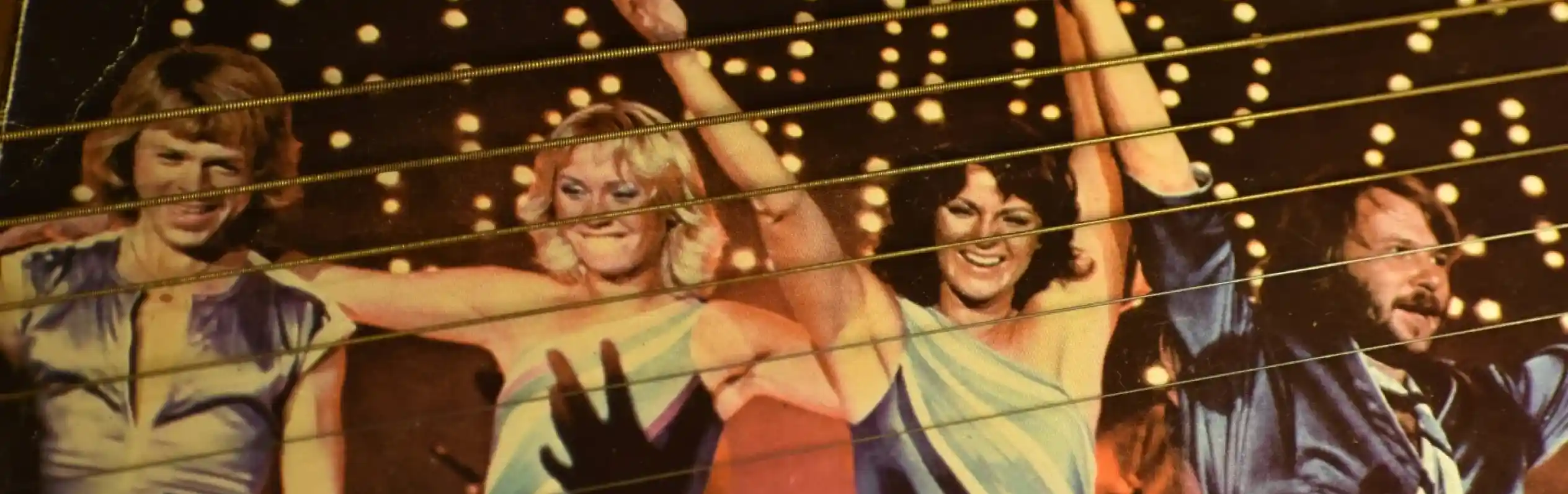 Four performers dressed in vibrant, sequined outfits pose with raised arms on stage, illuminated by bright lights. The crowd's hands can be seen reaching towards them in the foreground.