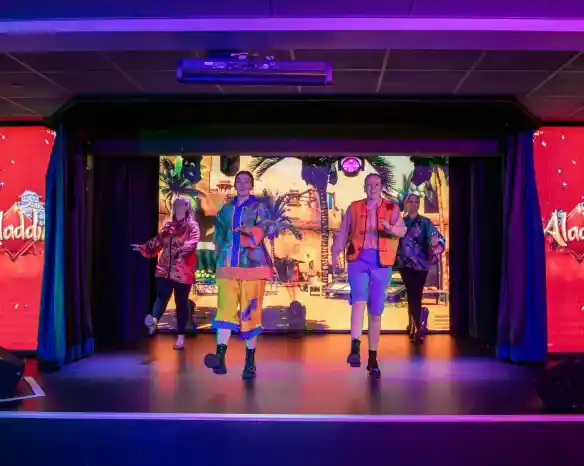 Four performers in colorful costumes dance on stage, with a vibrant backdrop featuring a scene from "Aladdin." The stage is lit with bold colors, creating an energetic atmosphere.