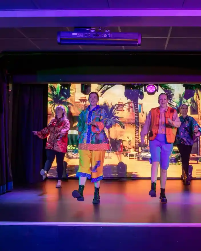 Four performers in colorful costumes dance on stage, with a vibrant backdrop featuring a scene from "Aladdin." The stage is lit with bold colors, creating an energetic atmosphere.