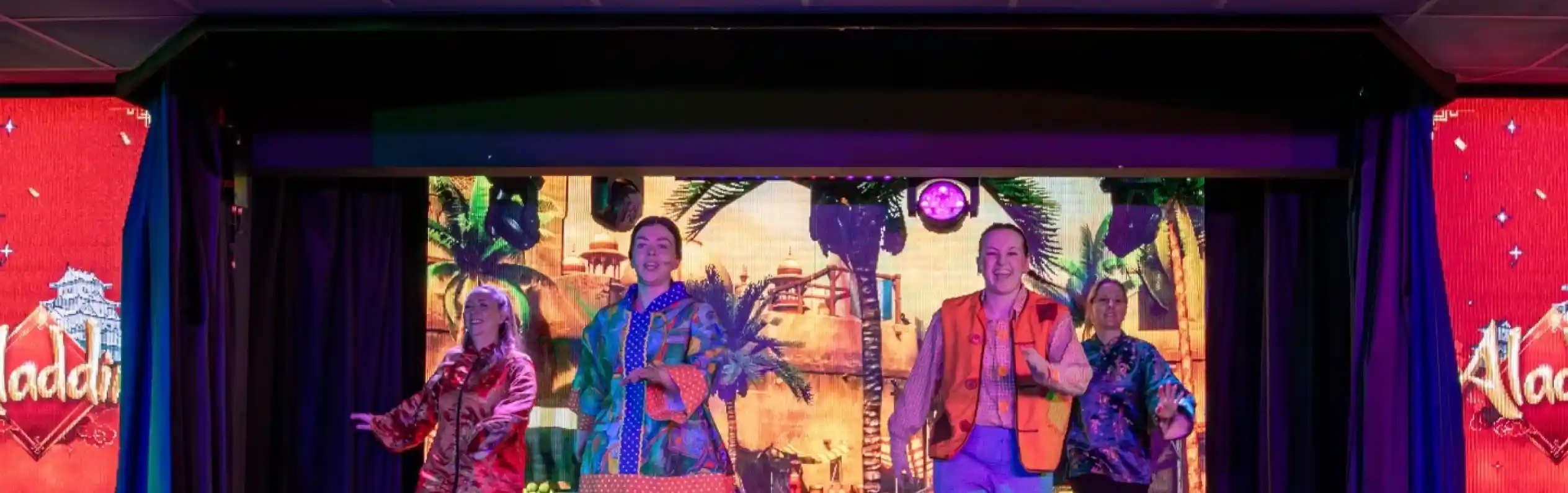 Four performers in colorful costumes dance on stage, with a vibrant backdrop featuring a scene from "Aladdin." The stage is lit with bold colors, creating an energetic atmosphere.