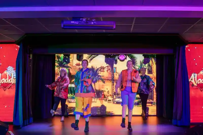 Four performers in colorful costumes dance on stage, with a vibrant backdrop featuring a scene from "Aladdin." The stage is lit with bold colors, creating an energetic atmosphere.