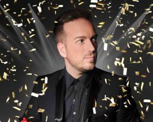 A man in a black suit with a black shirt holds a red object while looking off to the side. Gold confetti falls around him against a dark background with bright lights shining.