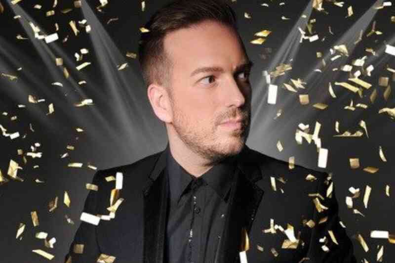 A man in a black suit with a black shirt holds a red object while looking off to the side. Gold confetti falls around him against a dark background with bright lights shining.