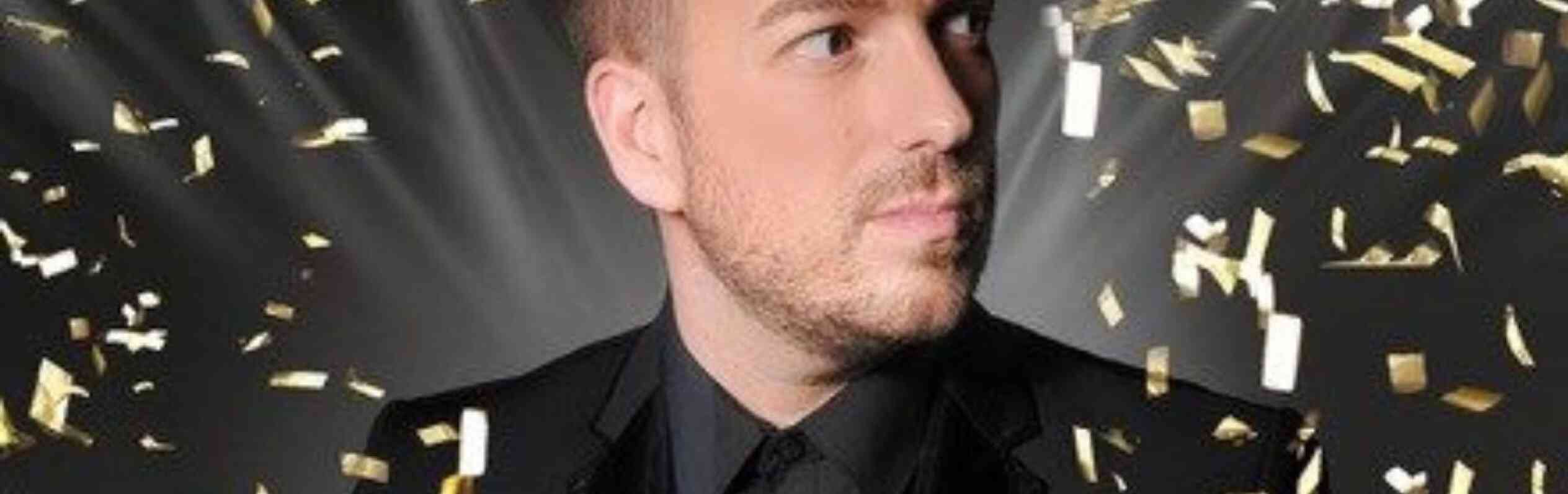 A man in a black suit with a black shirt holds a red object while looking off to the side. Gold confetti falls around him against a dark background with bright lights shining.