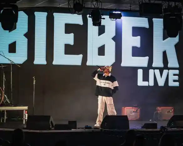 A performer on stage sings into a microphone, wearing a stylish outfit with a large graphic on their chest. Bright stage lights and a large screen in the background display the word "BIEBER" along with "LIVE." The audience can be seen in the foreground.