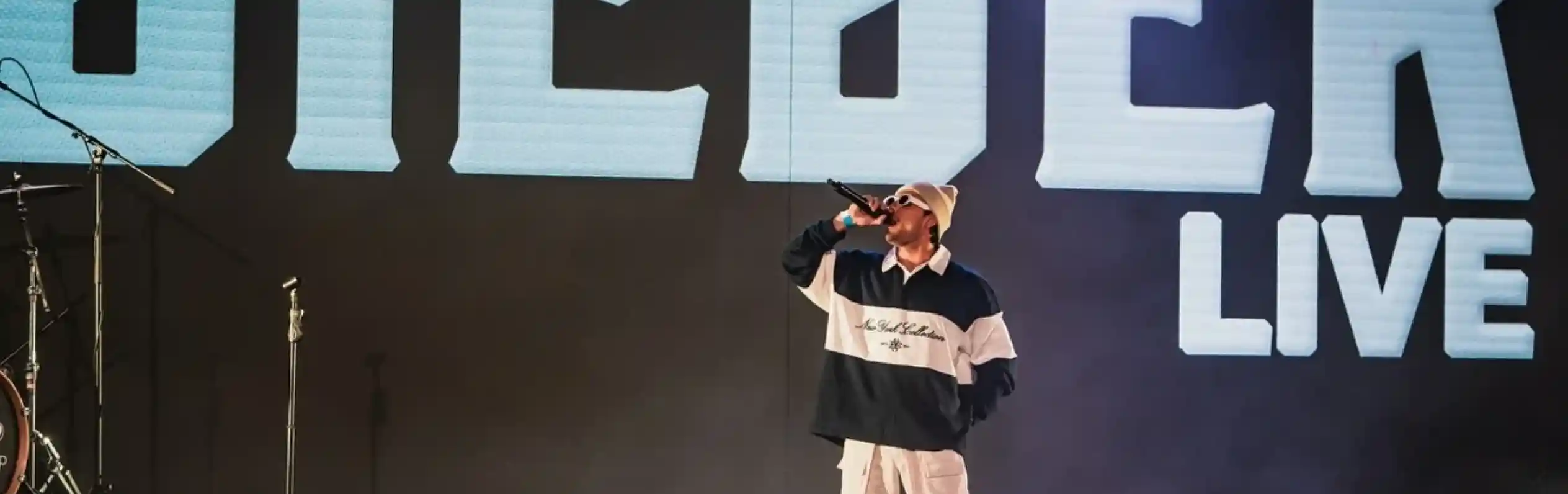 A performer on stage sings into a microphone, wearing a stylish outfit with a large graphic on their chest. Bright stage lights and a large screen in the background display the word "BIEBER" along with "LIVE." The audience can be seen in the foreground.