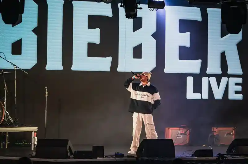 A performer on stage sings into a microphone, wearing a stylish outfit with a large graphic on their chest. Bright stage lights and a large screen in the background display the word "BIEBER" along with "LIVE." The audience can be seen in the foreground.