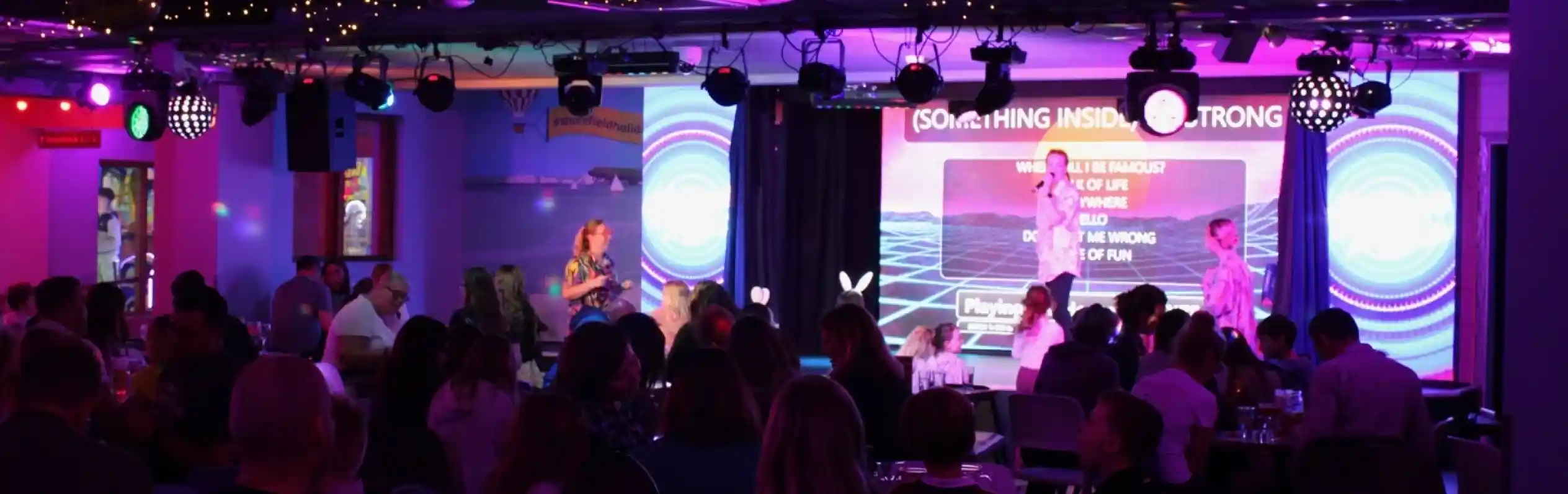 A lively entertainment venue filled with people enjoying a performance on stage, illuminated by colorful lights. Decorations include string lights and a backdrop with vibrant visuals. The audience, seated at tables, appears engaged and entertained.