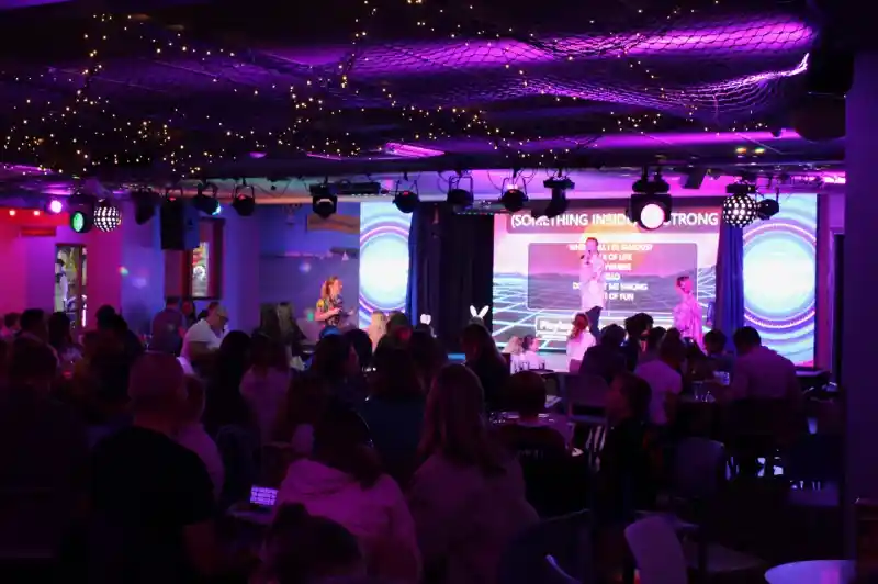 A lively entertainment venue filled with people enjoying a performance on stage, illuminated by colorful lights. Decorations include string lights and a backdrop with vibrant visuals. The audience, seated at tables, appears engaged and entertained.