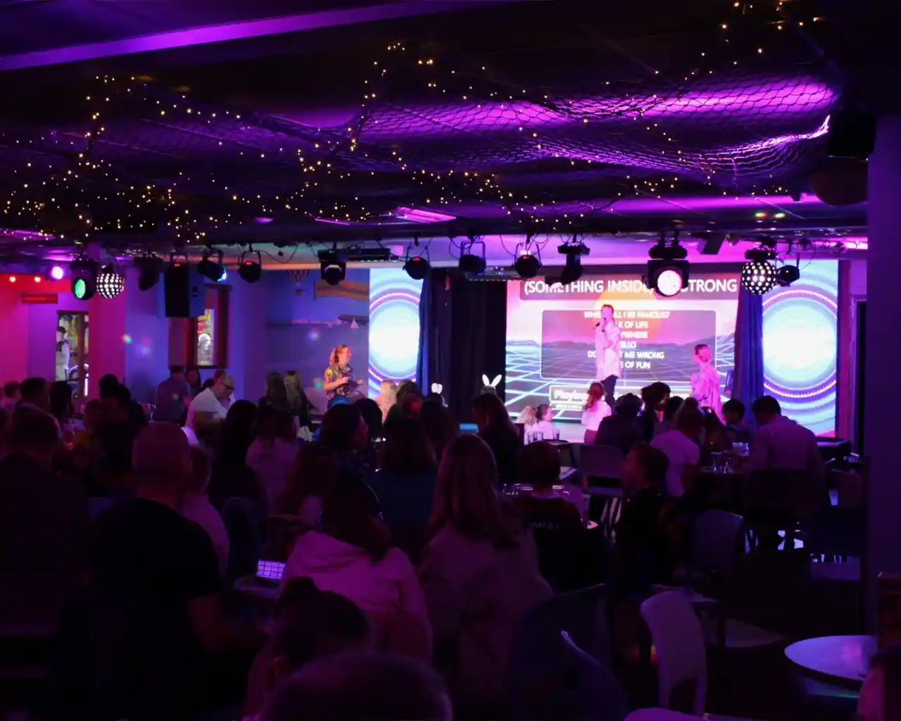 A lively entertainment venue filled with people enjoying a performance on stage, illuminated by colorful lights. Decorations include string lights and a backdrop with vibrant visuals. The audience, seated at tables, appears engaged and entertained.