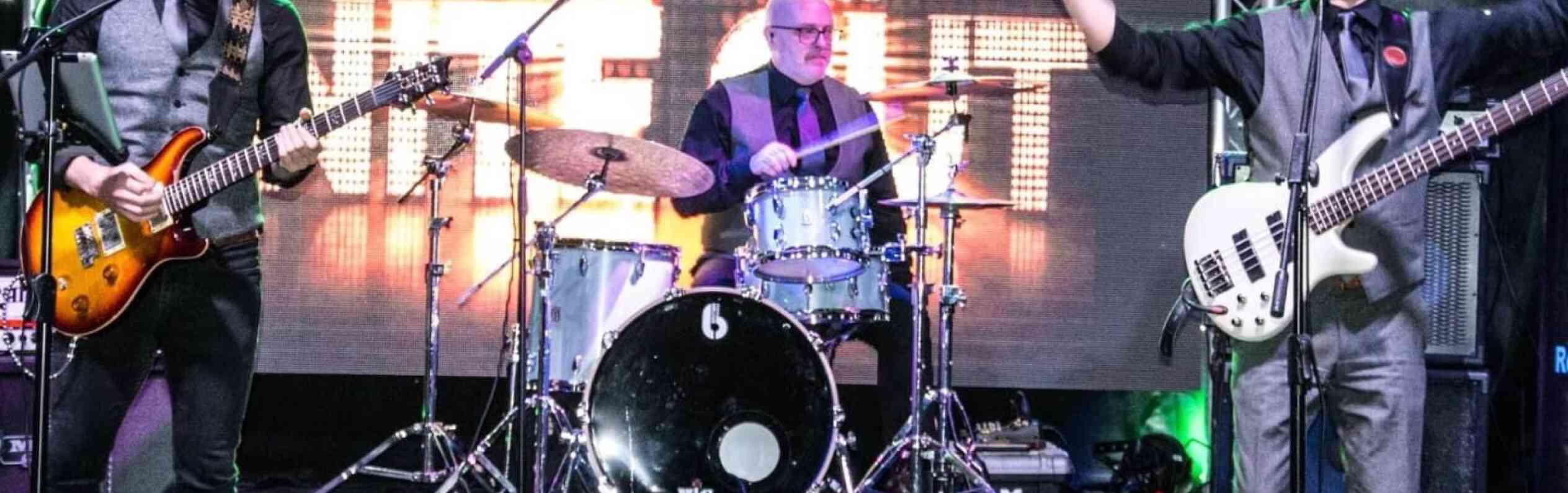 A live band performing on stage, featuring a guitarist and bassist on the left and right, with a drummer in the center. The backdrop displays "The Big Night Out" in bright letters, adding to the energetic atmosphere of the performance.