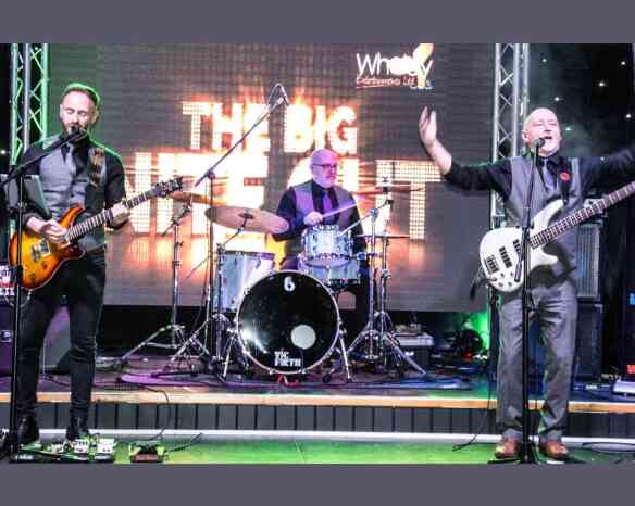 A live band performing on stage, featuring a guitarist and bassist on the left and right, with a drummer in the center. The backdrop displays "The Big Night Out" in bright letters, adding to the energetic atmosphere of the performance.