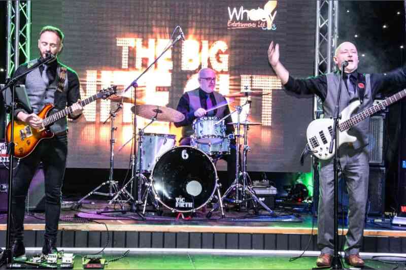 A live band performing on stage, featuring a guitarist and bassist on the left and right, with a drummer in the center. The backdrop displays "The Big Night Out" in bright letters, adding to the energetic atmosphere of the performance.
