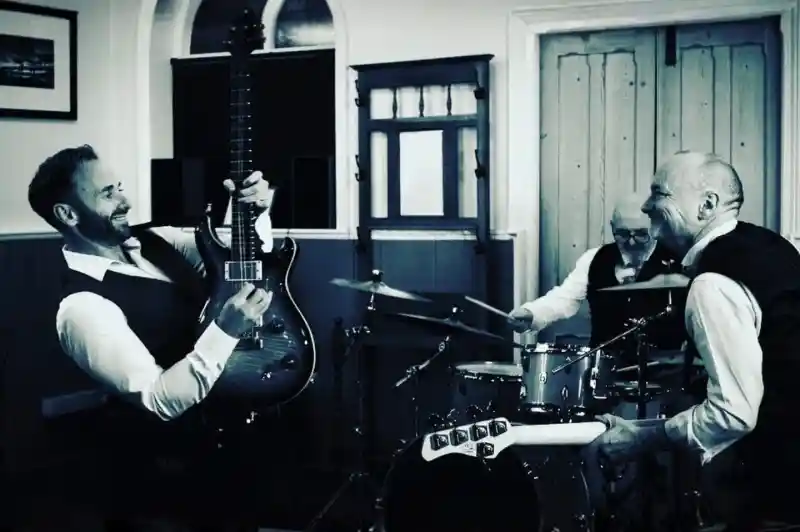 A black and white photo of three men playing music in a small venue. One man plays an electric guitar, smiling, while another plays drums in the background. The third man is singing and laughing. All are dressed in white shirts and black vests.