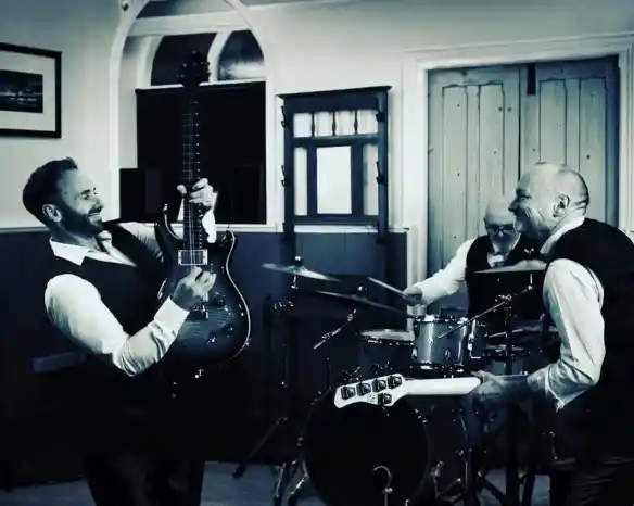 A black and white photo of three men playing music in a small venue. One man plays an electric guitar, smiling, while another plays drums in the background. The third man is singing and laughing. All are dressed in white shirts and black vests.