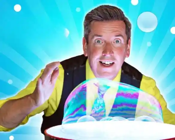 A cheerful man wearing a yellow shirt and a black vest enthusiastically interacts with a large soap bubble against a bright blue background with radiating lines.