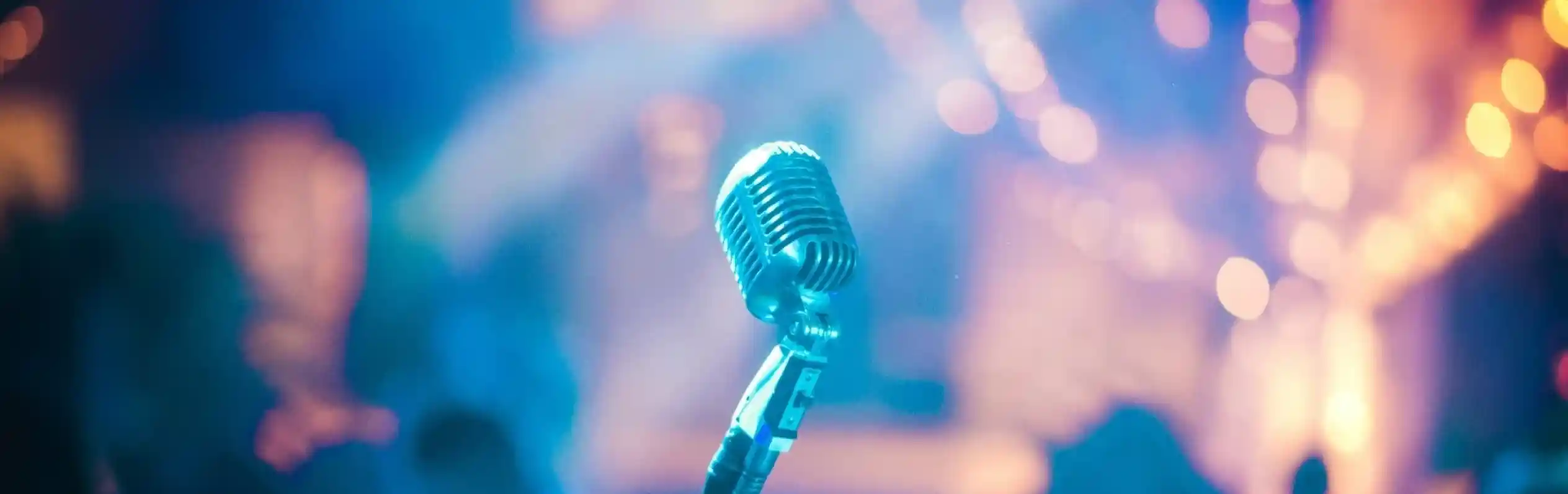 A vintage microphone stands in the foreground, surrounded by a blurred, colorful background with soft lights and silhouettes of people.