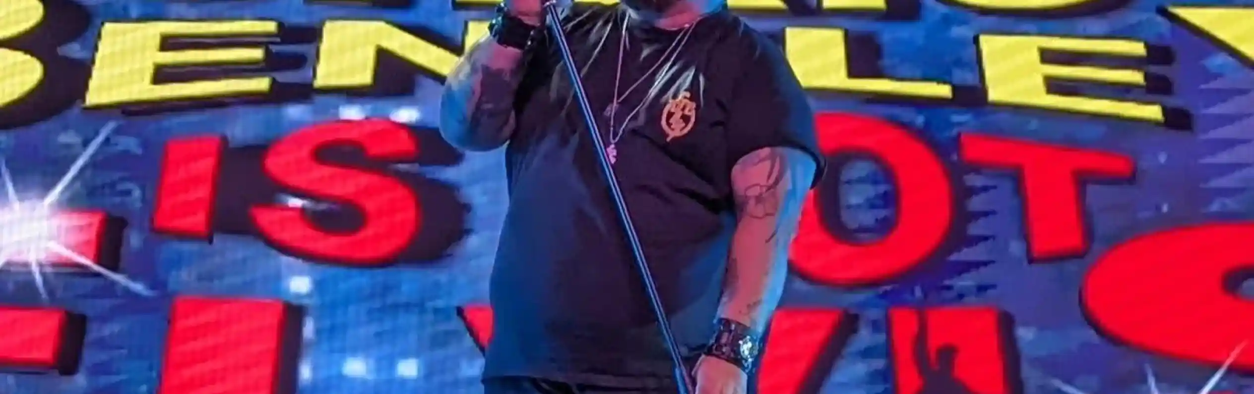 A performer, Chris Bentley, sings into a microphone on stage. Behind him is a vibrant backdrop featuring the text "Chris Bentley Is Not Elvis."