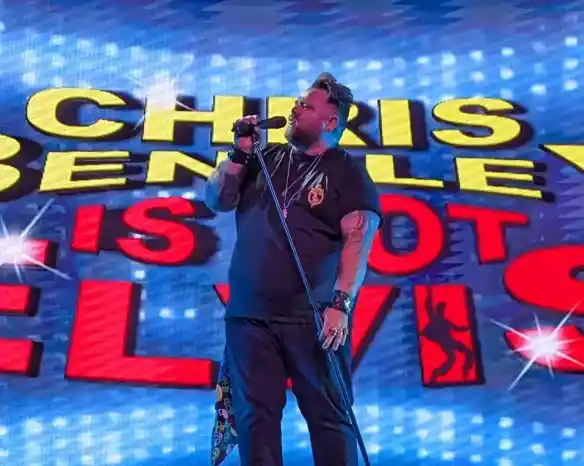 A performer, Chris Bentley, sings into a microphone on stage. Behind him is a vibrant backdrop featuring the text "Chris Bentley Is Not Elvis."
