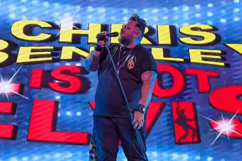 A performer, Chris Bentley, sings into a microphone on stage. Behind him is a vibrant backdrop featuring the text "Chris Bentley Is Not Elvis."