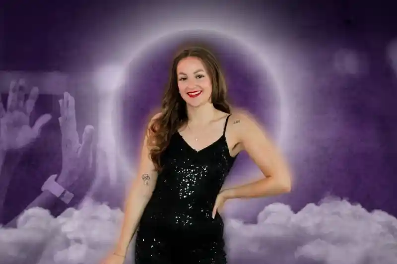 A smiling woman with long, wavy hair stands confidently in a sequined black jumpsuit. She poses in front of a purple and cloudy background, with raised hands in the foreground suggesting excitement or celebration.