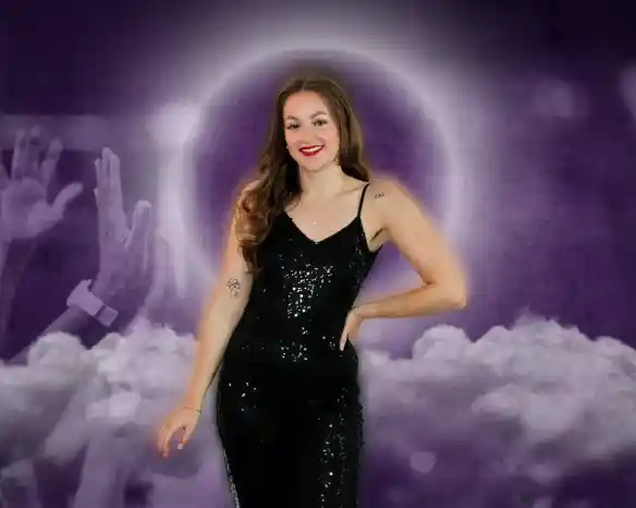 A smiling woman with long, wavy hair stands confidently in a sequined black jumpsuit. She poses in front of a purple and cloudy background, with raised hands in the foreground suggesting excitement or celebration.