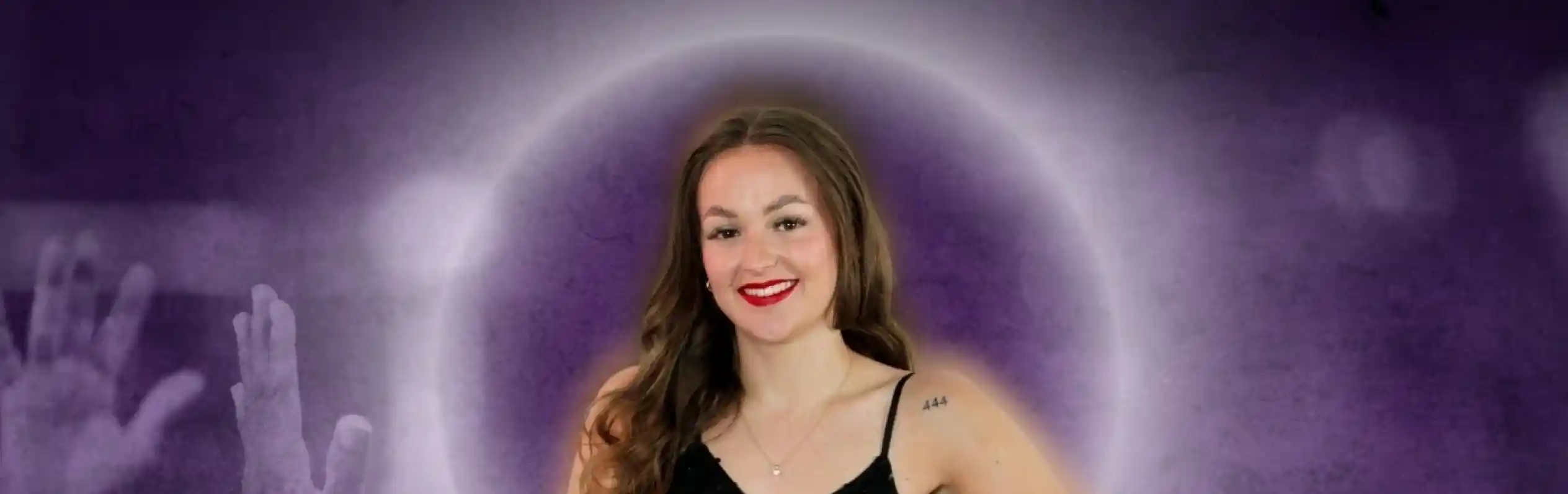 A smiling woman with long, wavy hair stands confidently in a sequined black jumpsuit. She poses in front of a purple and cloudy background, with raised hands in the foreground suggesting excitement or celebration.