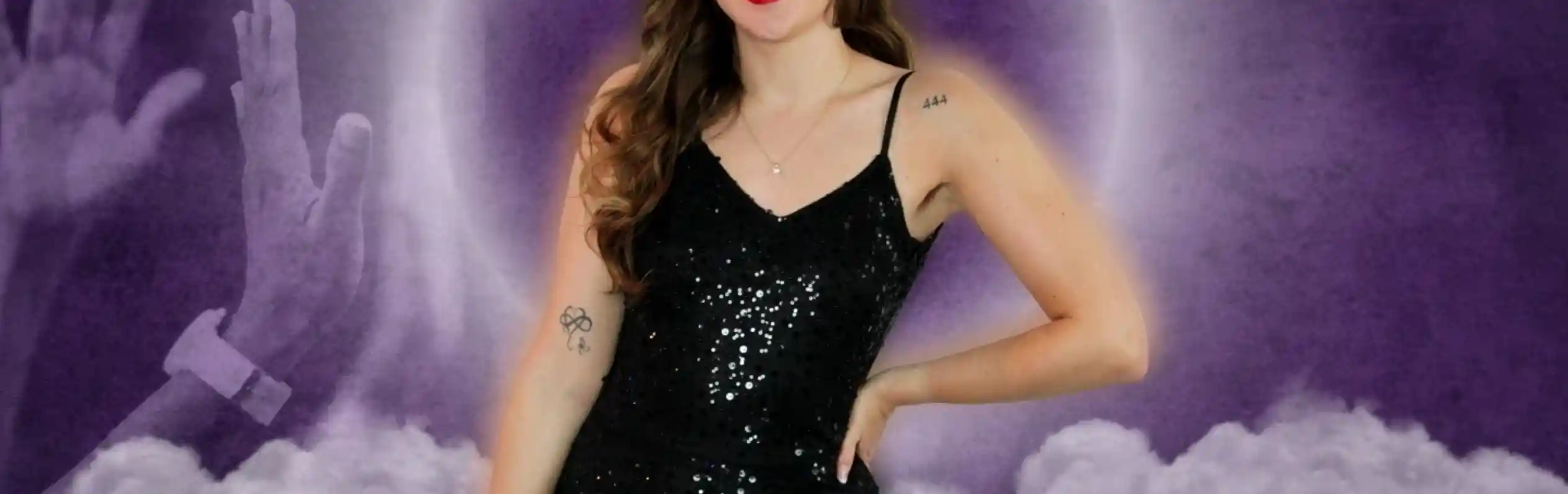 A smiling woman with long, wavy hair stands confidently in a sequined black jumpsuit. She poses in front of a purple and cloudy background, with raised hands in the foreground suggesting excitement or celebration.