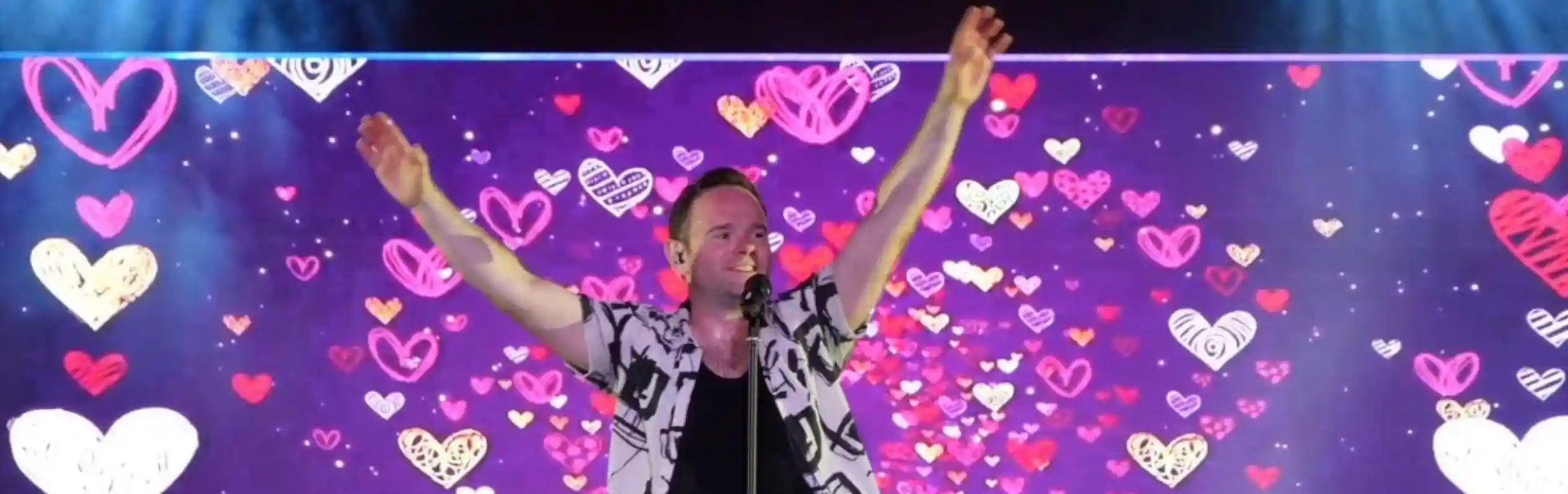 A performer stands on stage with arms raised, smiling and engaging with the audience. A colorful backdrop features a pattern of hearts in various shades. Fans in the foreground raise their hands in excitement.