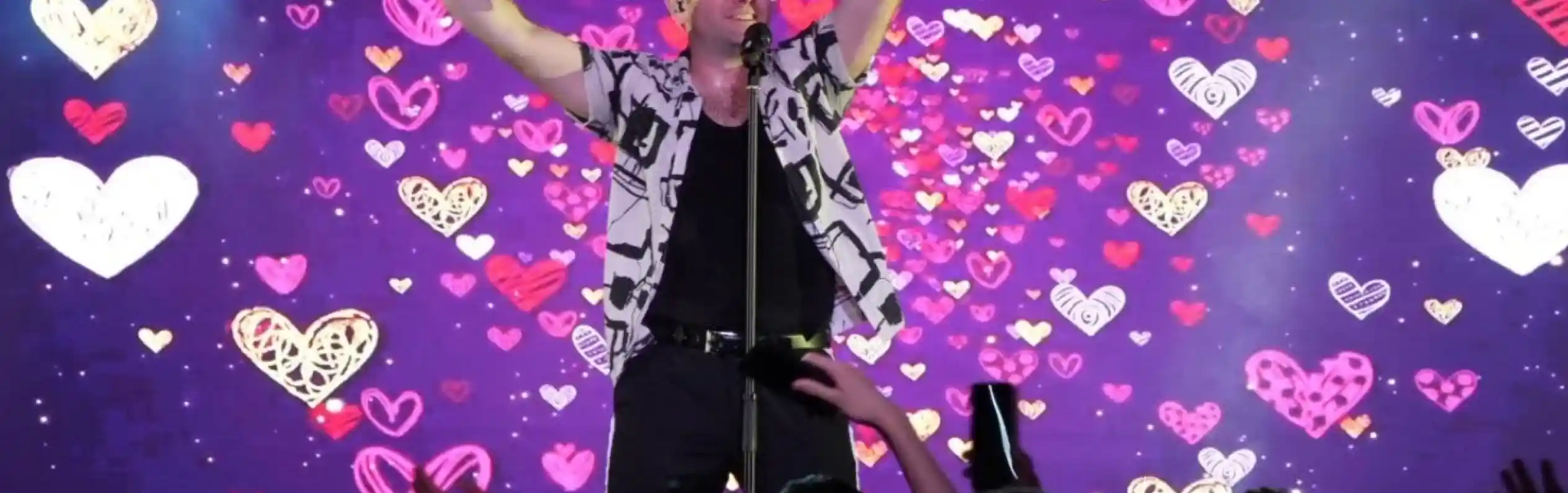 A performer stands on stage with arms raised, smiling and engaging with the audience. A colorful backdrop features a pattern of hearts in various shades. Fans in the foreground raise their hands in excitement.