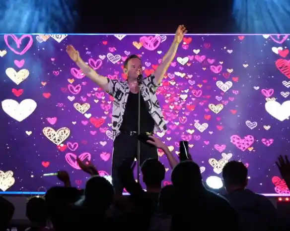 A performer stands on stage with arms raised, smiling and engaging with the audience. A colorful backdrop features a pattern of hearts in various shades. Fans in the foreground raise their hands in excitement.