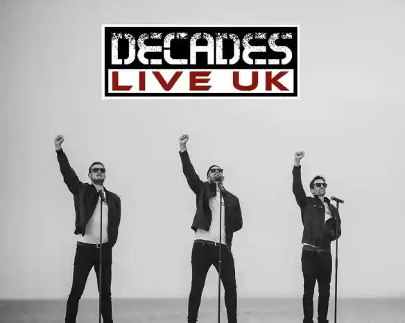 Three singers stand on a beach with raised fists, wearing sunglasses and black leather jackets. They are positioned in front of microphones, creating a dynamic performance vibe. The background features a grey sky and ocean. The logo "Decades Live UK" is displayed at the top.