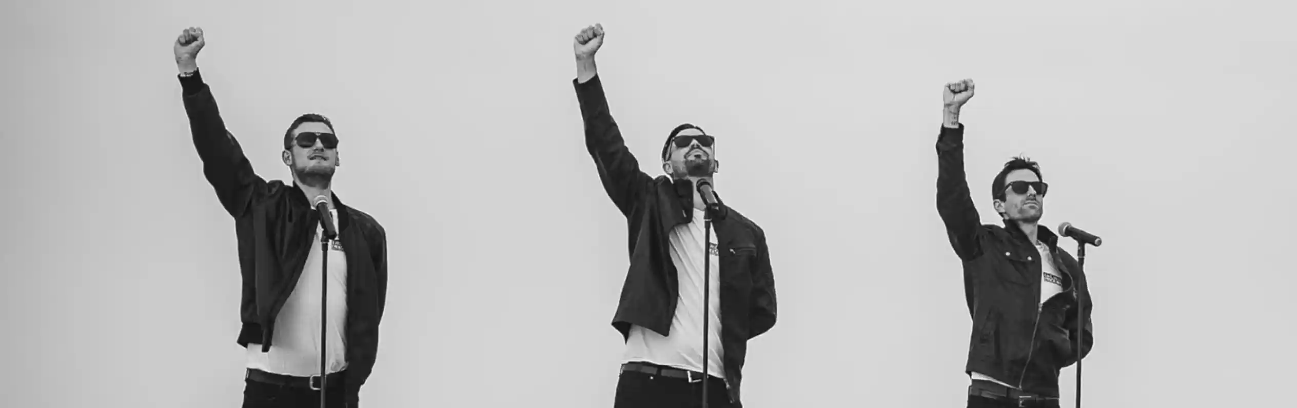 Three singers stand on a beach with raised fists, wearing sunglasses and black leather jackets. They are positioned in front of microphones, creating a dynamic performance vibe. The background features a grey sky and ocean. The logo "Decades Live UK" is displayed at the top.