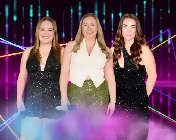Three women stand side by side, each wearing sparkly outfits. The first woman has long, straight red hair and is in a black dress. The second woman, with long blonde hair, wears a white top and shiny green pants. The third woman has long, wavy dark hair and is dressed in a black, sparkly outfit. The background features colorful lights and patterns creating a vibrant atmosphere.