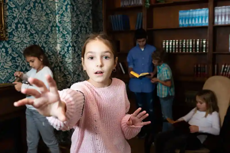 Group of children and young teens solving puzzles in a themed escape room, with one girl in a pink sweater reaching toward the camera while others search for clues among bookshelves and furniture.