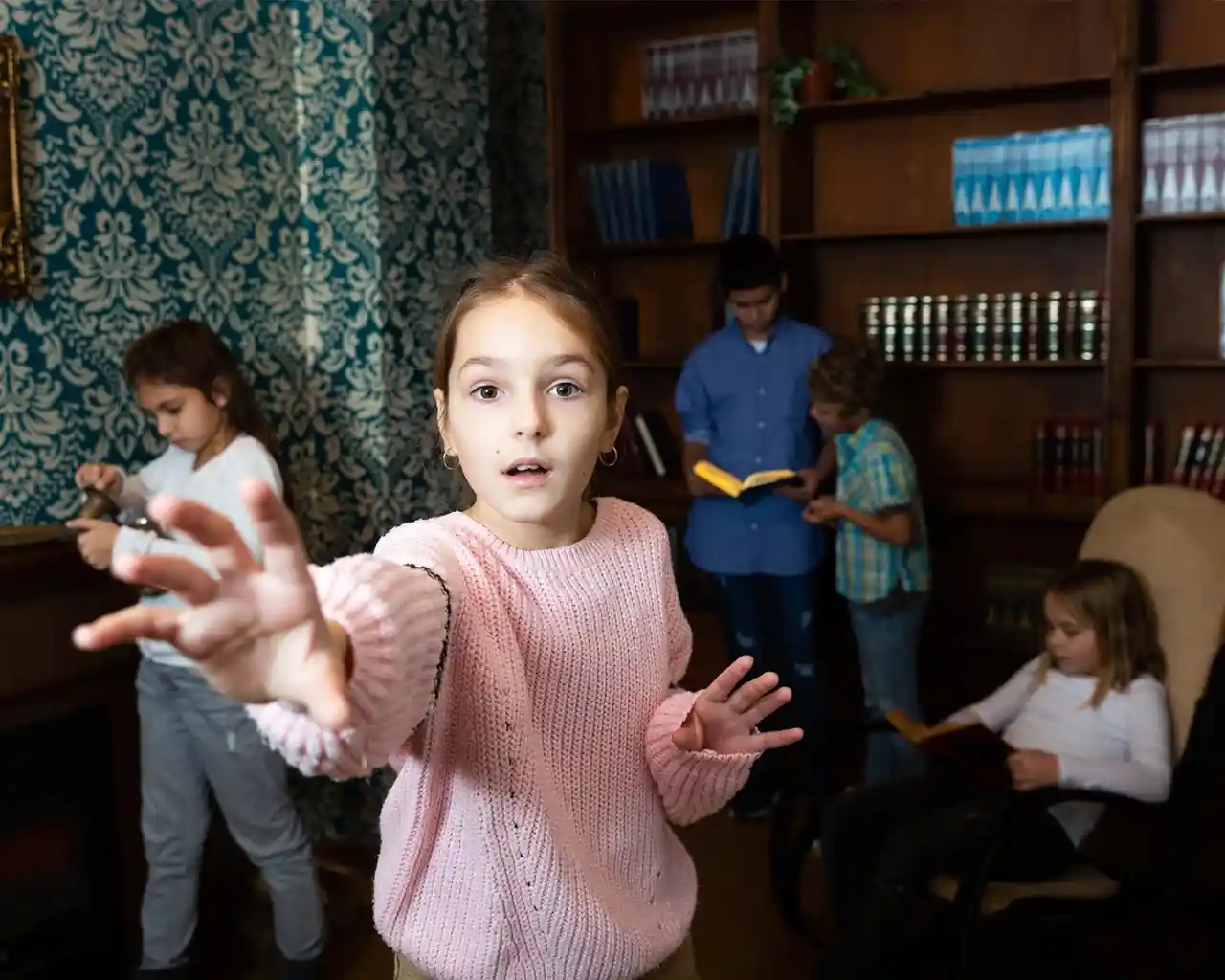 Group of children and young teens solving puzzles in a themed escape room, with one girl in a pink sweater reaching toward the camera while others search for clues among bookshelves and furniture.