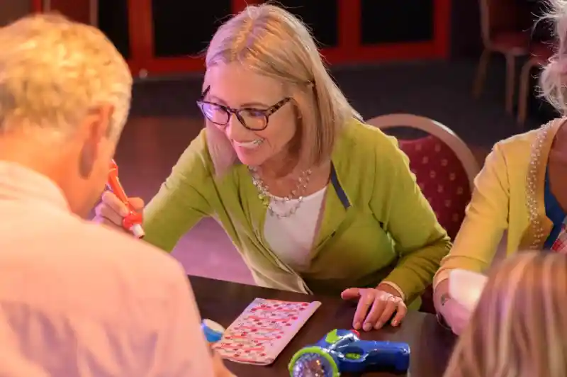 A smiling woman marks her bingo card with a dabber while sitting at a table with other players during a lively bingo session.