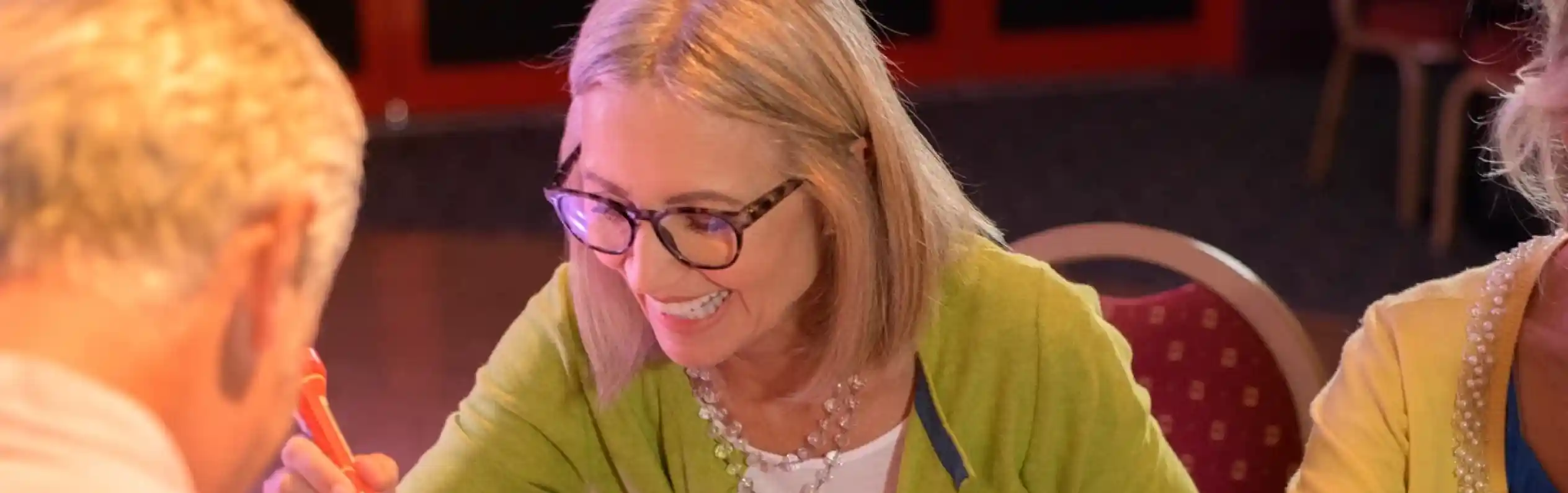 A smiling woman marks her bingo card with a dabber while sitting at a table with other players during a lively bingo session.