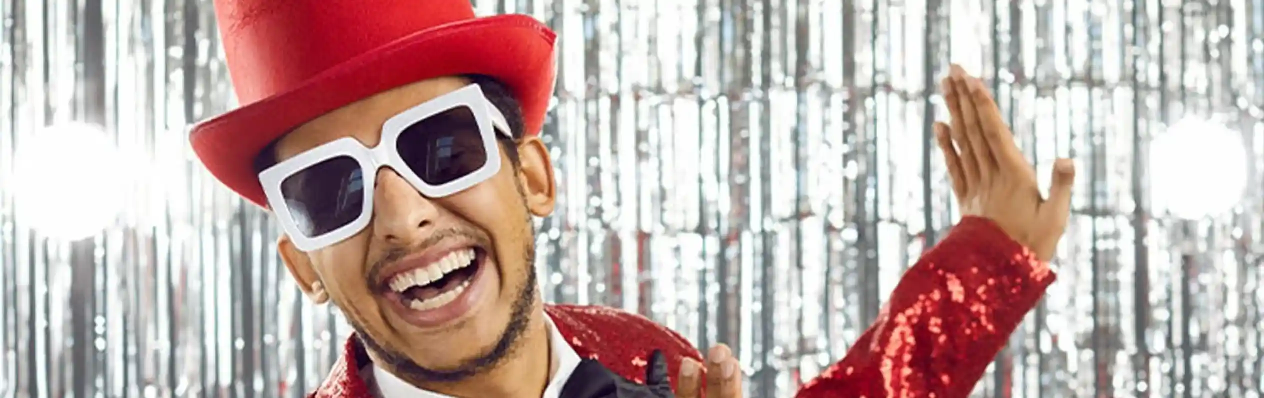 A smiling man wearing a red sequined suit, a black bow tie, and oversized white sunglasses. He is striking a playful pose in front of a shiny silver backdrop.