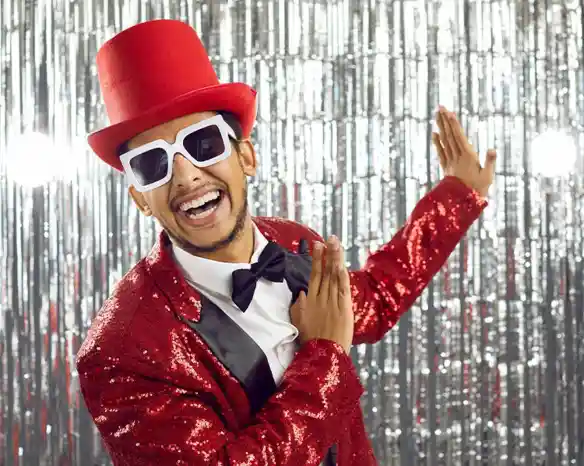 A smiling man wearing a red sequined suit, a black bow tie, and oversized white sunglasses. He is striking a playful pose in front of a shiny silver backdrop.