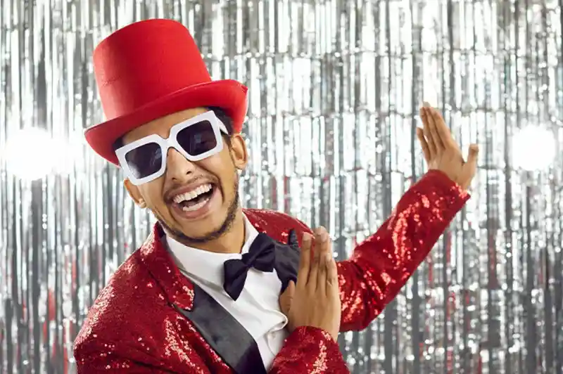 A smiling man wearing a red sequined suit, a black bow tie, and oversized white sunglasses. He is striking a playful pose in front of a shiny silver backdrop.