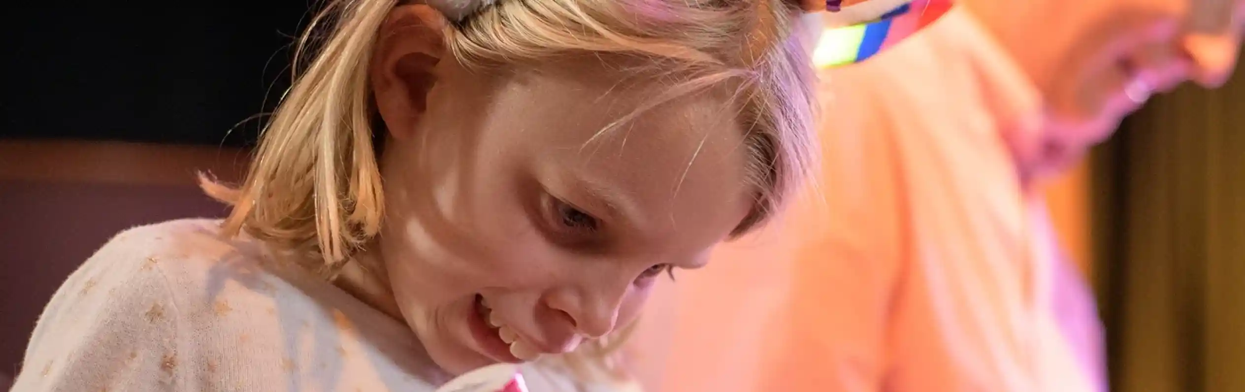 A young girl wearing a sparkly unicorn headband and star-patterned top excitedly marks her bingo card with a pink dabber. She is focused and smiling, sitting at a table next to an older adult also playing bingo.