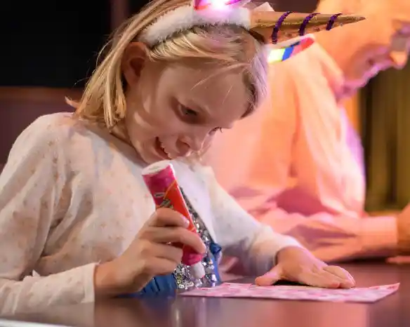 A young girl wearing a sparkly unicorn headband and star-patterned top excitedly marks her bingo card with a pink dabber. She is focused and smiling, sitting at a table next to an older adult also playing bingo.