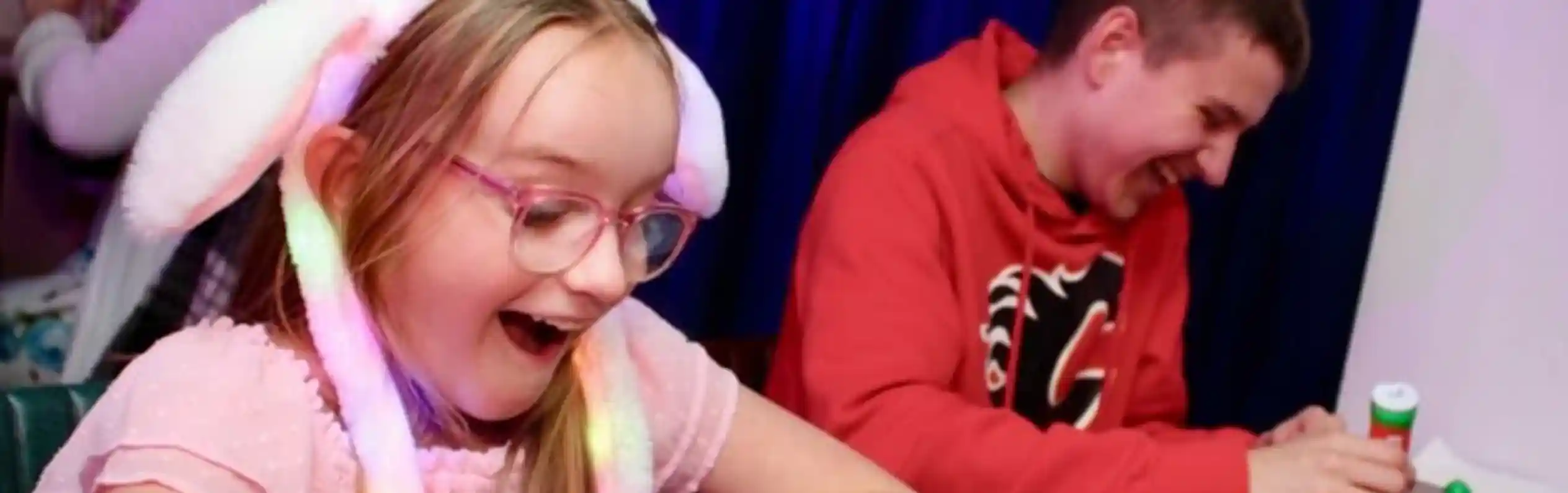 A young girl wearing a light-up bunny ear hat and pink dress smiles with excitement while playing bingo at a table. Next to her, a teenage boy in a red hoodie laughs as he marks his bingo card. Colorful bingo dabbers and balls are on the table, with a dark blue curtain in the background and other people playing in the distance.