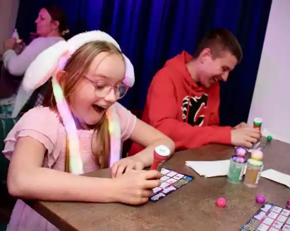 A young girl wearing a light-up bunny ear hat and pink dress smiles with excitement while playing bingo at a table. Next to her, a teenage boy in a red hoodie laughs as he marks his bingo card. Colorful bingo dabbers and balls are on the table, with a dark blue curtain in the background and other people playing in the distance.