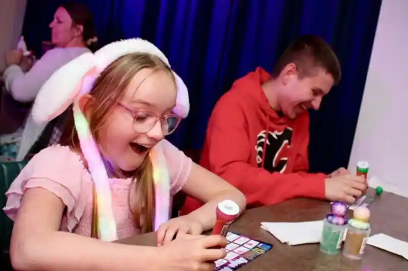 A young girl wearing a light-up bunny ear hat and pink dress smiles with excitement while playing bingo at a table. Next to her, a teenage boy in a red hoodie laughs as he marks his bingo card. Colorful bingo dabbers and balls are on the table, with a dark blue curtain in the background and other people playing in the distance.