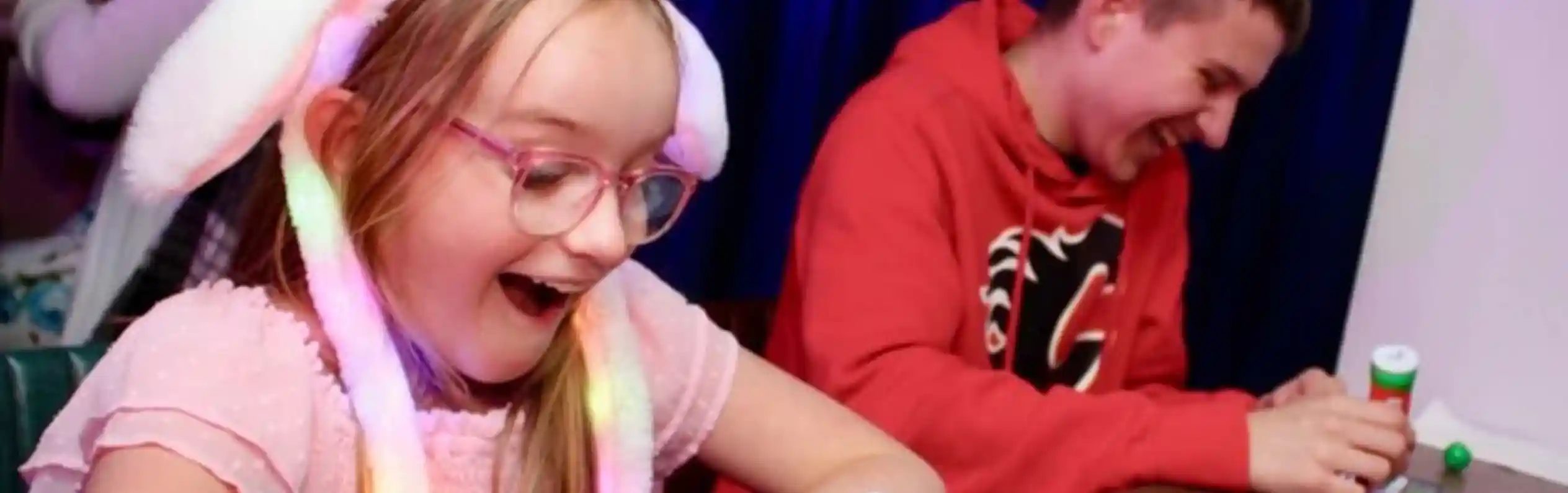A young girl wearing a light-up bunny ear hat and pink dress smiles with excitement while playing bingo at a table. Next to her, a teenage boy in a red hoodie laughs as he marks his bingo card. Colorful bingo dabbers and balls are on the table, with a dark blue curtain in the background and other people playing in the distance.