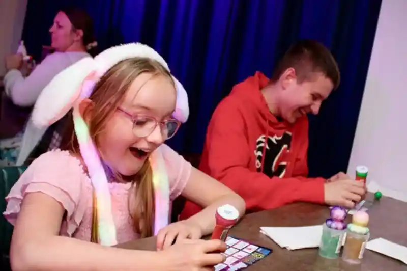 A young girl wearing a light-up bunny ear hat and pink dress smiles with excitement while playing bingo at a table. Next to her, a teenage boy in a red hoodie laughs as he marks his bingo card. Colorful bingo dabbers and balls are on the table, with a dark blue curtain in the background and other people playing in the distance.