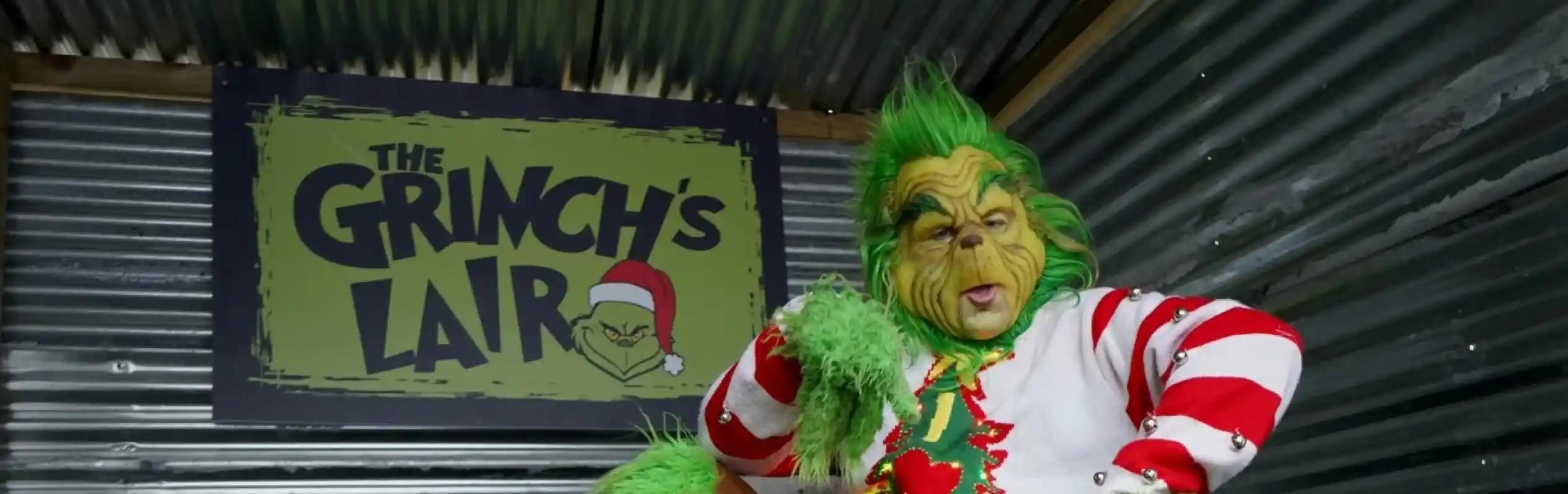 The Grinch, dressed in festive attire with green fur and a mischievous expression, sits in a wooden chair in front of a sign that reads "The Grinch's Lair." Decorated with holiday themes, the setting includes a corrugated metal backdrop and a small evergreen in the corner.